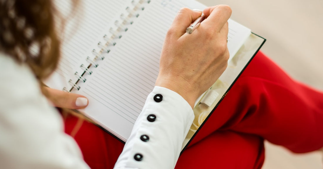 A woman writing notes by hand in an open spiral notebook, seen over her shoulder