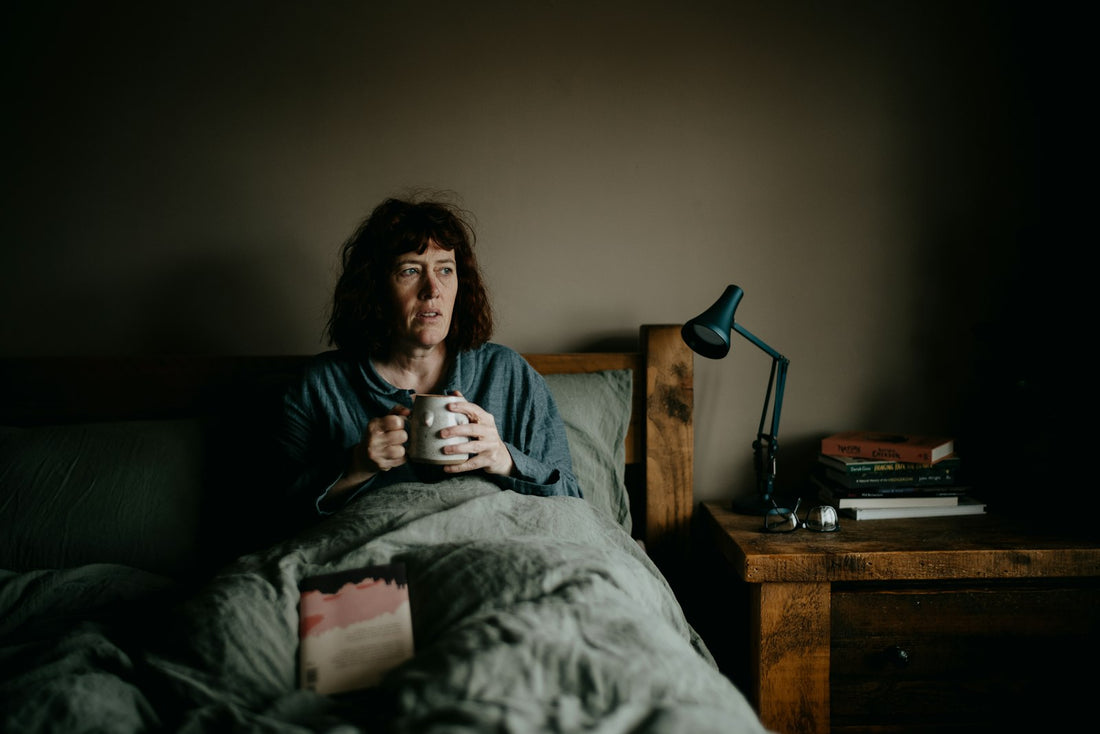 A woman sitting up in bed holding a mug, with a desk lamp and books on the bedside table