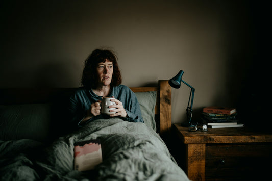 A woman sitting up in bed holding a mug, with a desk lamp and books on the bedside table