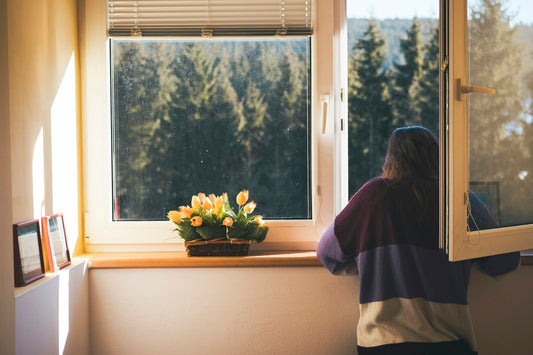 A woman in a striped jumper looking out of an open window at pine trees, with yellow tulips on the sill in morning sun