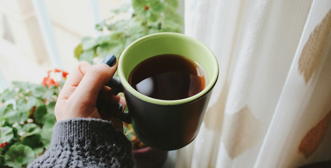 A hand in a knitted jumper sleeve holding a mug of tea by a window