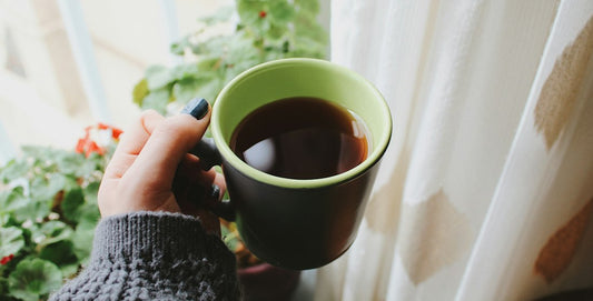 A hand in a knitted jumper sleeve holding a mug of tea by a window