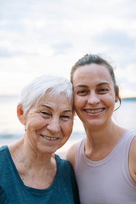 An older woman with short white hair and her adult daughter smiling cheek to cheek by the sea