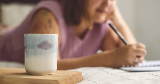 A woman writing in a notebook at a table at home, a mug beside her, soft daylight