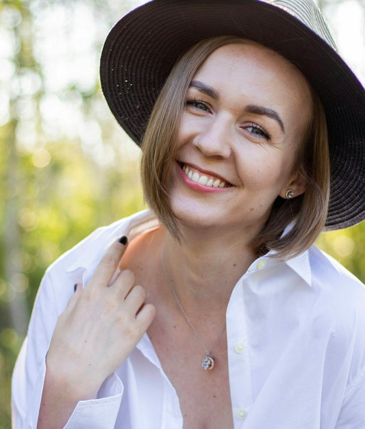 A smiling woman in a wide-brimmed hat and white shirt outdoors in soft sunlight