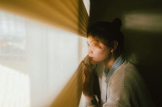 A woman in a cardigan looking out through a curtain at early morning light