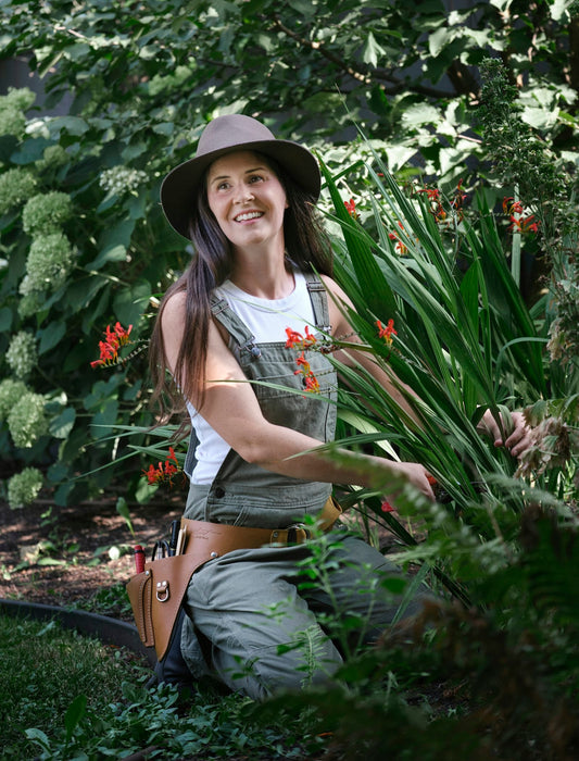 A smiling woman in a brown hat and dungarees kneeling among green plants and orange flowers in a garden