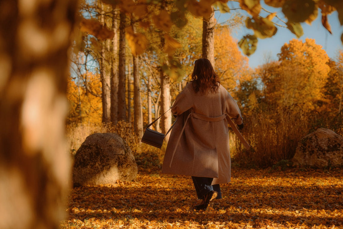 A woman in a long camel coat walking through an autumn forest with golden leaves