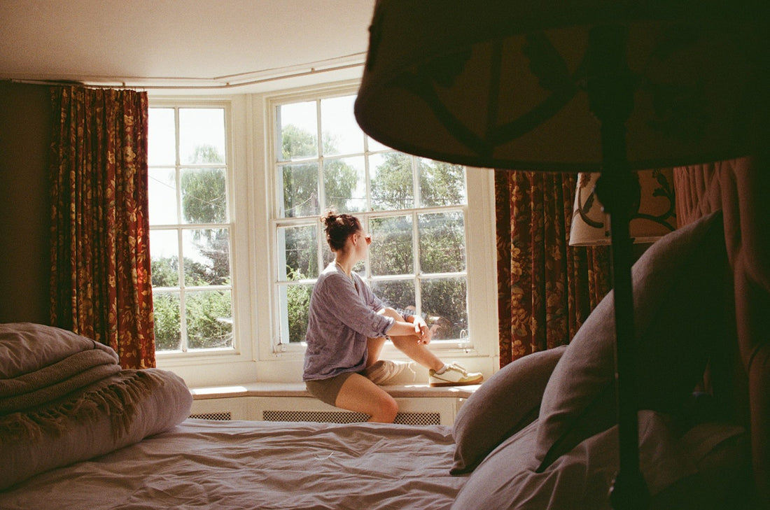 A woman sitting on a window seat in a sunlit bay window, looking out at the garden