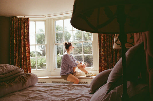 A woman sitting on a window seat in a sunlit bay window, looking out at the garden
