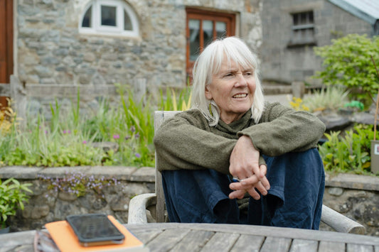 A smiling white-haired woman sitting relaxed at a wooden garden table in front of a stone house